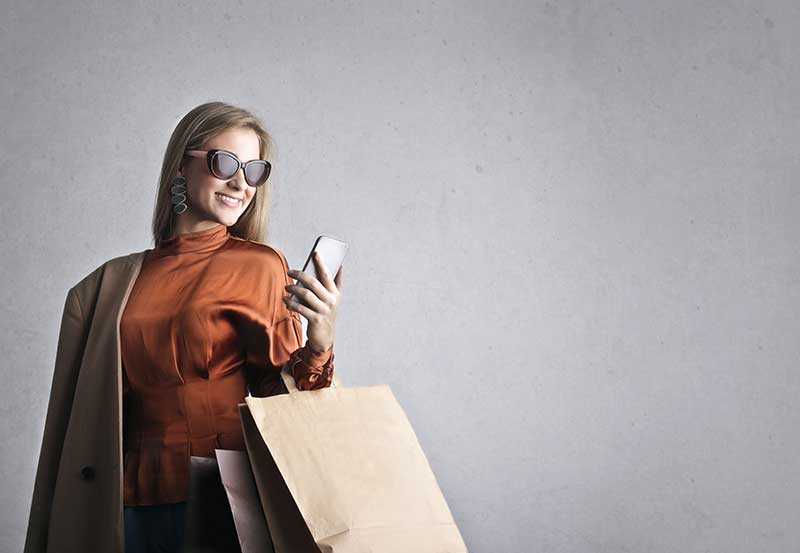 A stylish woman wearing sunglasses holds shopping bags and a phone, representing boutique fashion online shopping.